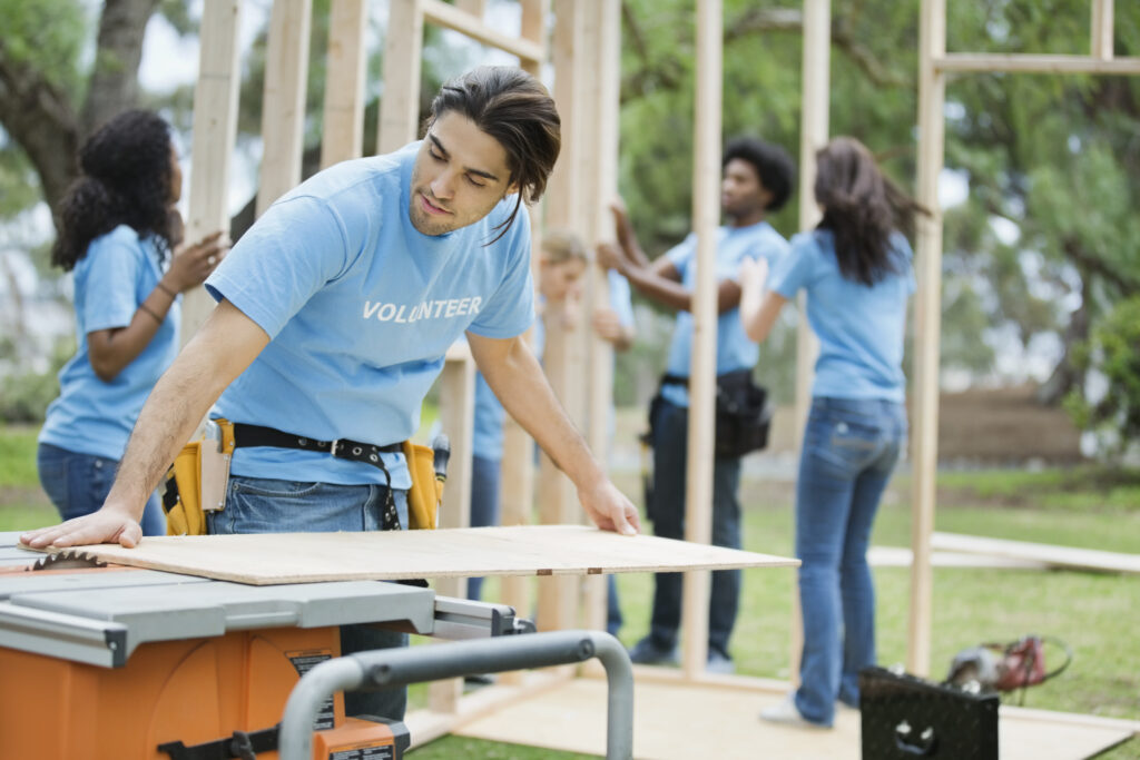 Young man cutting wood with table saw and volunteers building fr